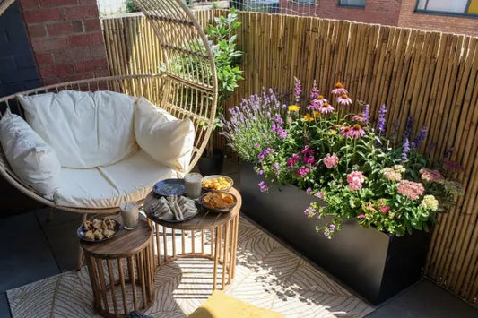 Cosy small balcony with a wicker egg chair and cream cushions beside a bamboo privacy screen, a long black planter overflowing with colourful flowers