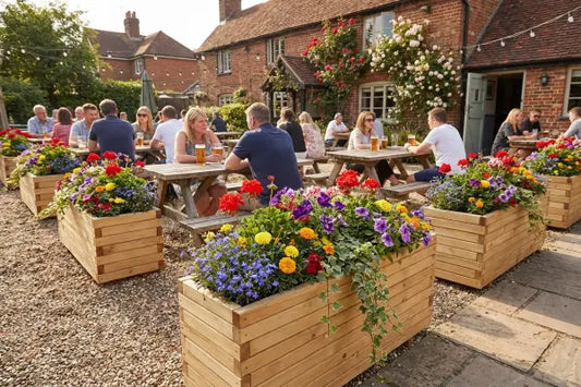 People relax at wooden picnic tables in a sunny pub garden, drinking pints among colourful flower planters and string lights beside a red-brick building with climbing roses.