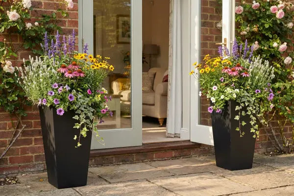 Two tall black planters overflowing with colourful flowers and trailing ivy flank open French doors on a brick house, with climbing roses around the doorway, a sunlit stone patio, and a cosy sofa visible inside.