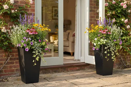 Two tall black planters overflowing with colourful flowers and trailing ivy flank open French doors on a brick house, with climbing roses around the doorway, a sunlit stone patio, and a cosy sofa visible inside.
