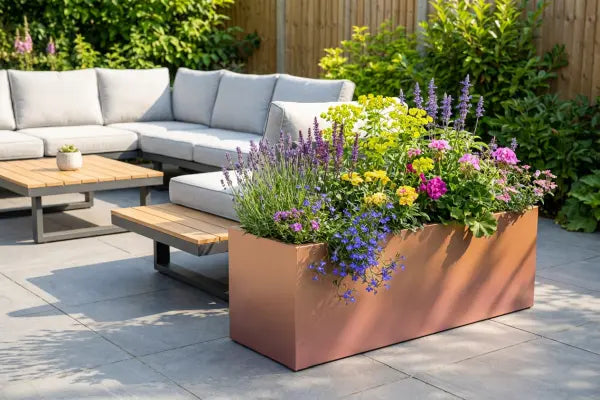 Sunny backyard patio with a gray cushioned sectional and wood-top coffee tables beside a large rectangular planter overflowing with colourful flowers