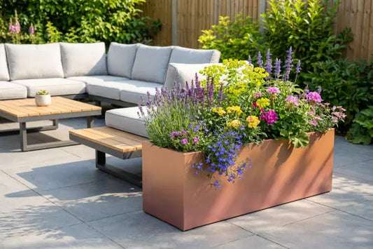 Sunny backyard patio with a gray cushioned sectional and wood-top coffee tables beside a large rectangular planter overflowing with colourful flowers