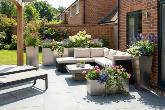 Sunny backyard patio beside a brick house featuring an L‑shaped cream‑cushioned wicker sofa and low concrete table, surrounded by sleek silver planters overflowing with colourful flowers