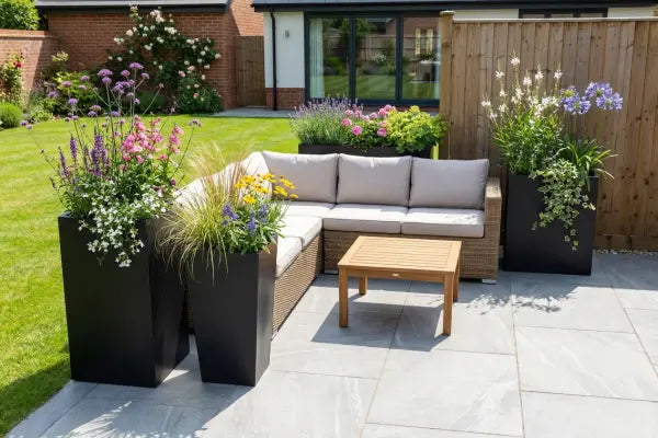 Sunny backyard patio with an L-shaped wicker sofa and small wooden coffee table on gray stone tiles, bordered by tall black planters bursting with colourful flowers and grasses, with a wooden fence, green lawn, and modern brick house in the background.