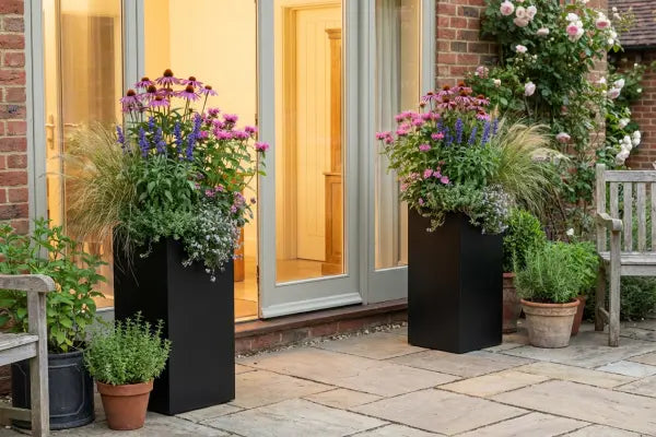 Two tall black planters overflowing with pink and purple flowers and feathery grasses flank glass patio doors of a brick house, surrounded by terracotta herb pots, a wooden bench, and climbing roses, with warm light glowing from inside.