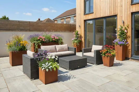 Modern backyard patio featuring a wicker sofa set with beige cushions and a low table on pale stone paving, encircled by Corten planters overflowing with colourful flowers and grasses
