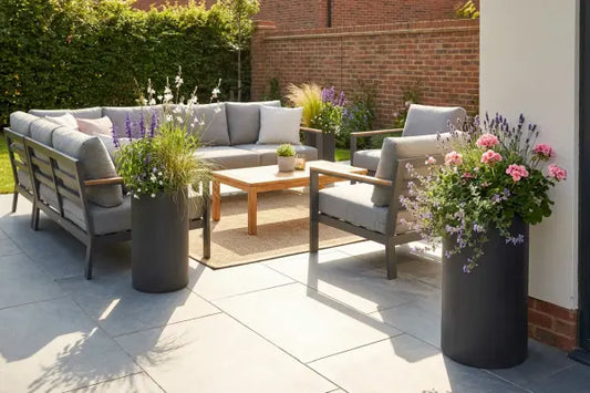 Sunny backyard patio with gray cushioned sectional and armchairs around a wooden coffee table on a rug, set on large stone pavers and framed by tall black planters