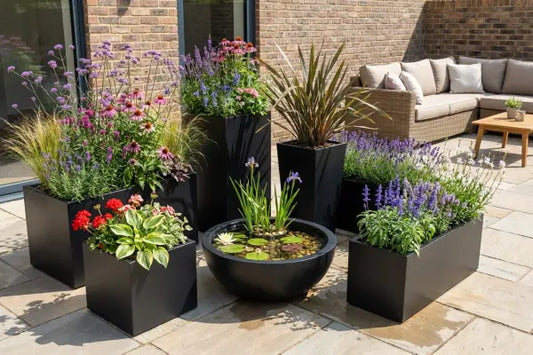 Modern patio with sleek black planters filled with colourful flowers and grasses surrounding a round bowl water feature with lilies and irises, set on stone paving beside a brick wall with a rattan sectional sofa and small wooden table.