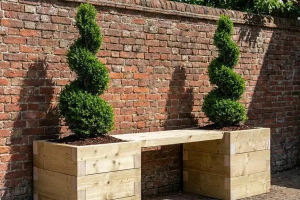 A light-wood bench bridges two square planters with spiral-trimmed topiary shrubs, set against a weathered red-brick wall in a sunny garden courtyard.