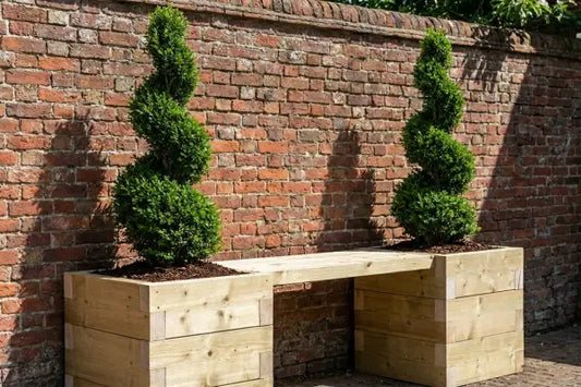 A light-wood bench bridges two square planters with spiral-trimmed topiary shrubs, set against a weathered red-brick wall in a sunny garden courtyard.