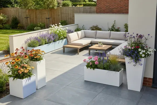 Modern backyard patio with an L-shaped beige outdoor sofa and wooden coffee tables on gray pavers, bordered by white planters bursting with colourful flowers and grasses