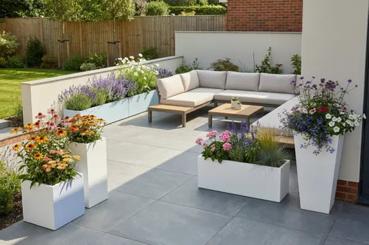 Modern backyard patio with an L-shaped beige outdoor sofa and wooden coffee tables on gray pavers, bordered by white planters bursting with colourful flowers and grasses