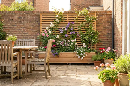 Sunny brick-walled patio featuring a weathered wooden table and chairs beside a slatted trellis planter covered in pink and white roses, purple clematis, and other blooms, with terracotta pots of geraniums and herbs scattered across the paving.