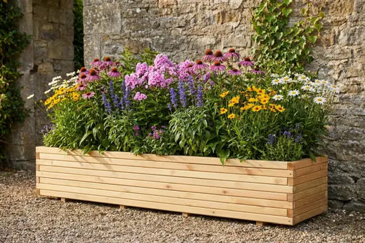 A long light-wood planter overflows with colourful summer flowers—pink coneflowers, purple spikes, yellow blooms, and white daisies—set on a gravel surface against a rustic stone wall with trailing ivy.